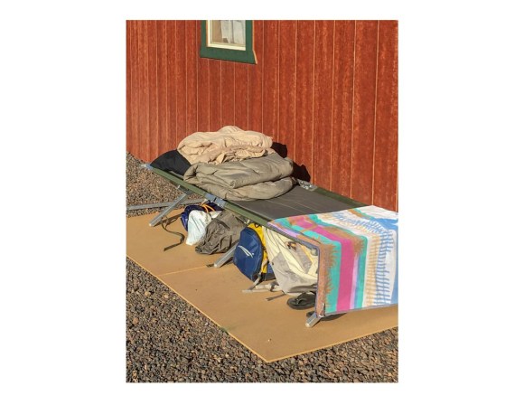 Sleeping cot with bedding on top and duffle bag and other camping gear underneath, set up next to wood-paneled building.