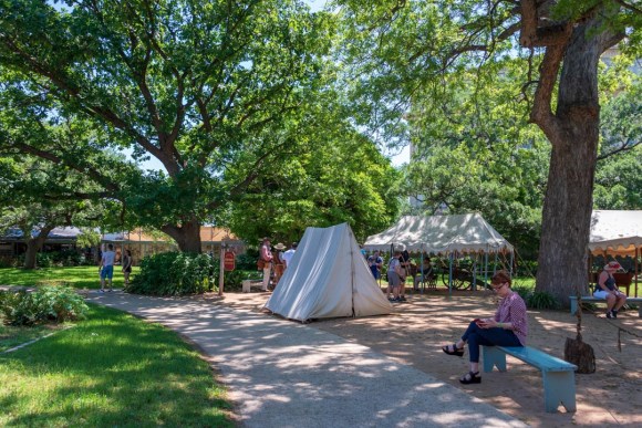 Tent used in reenactment of Alamo history, with visitors on landscaped grounds in background.