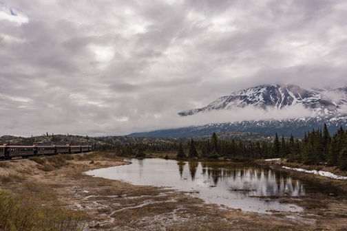 Landscape image of snow-capped mountains, cloudy grey skies, small pond next to evergreen trees, and train cars in the background.