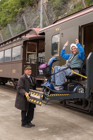 Train conductor using hydraulic lift to transport woman in mobility scooter out of train car.