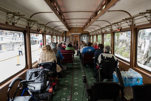 View, from rear, of passengers in wheelchairs, mobility scooters or train seats inside train car prior to leaving station.