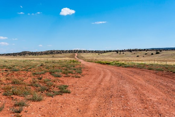 Landscape image of red dirt road in foreground, with small hills and vast blue sky in background.