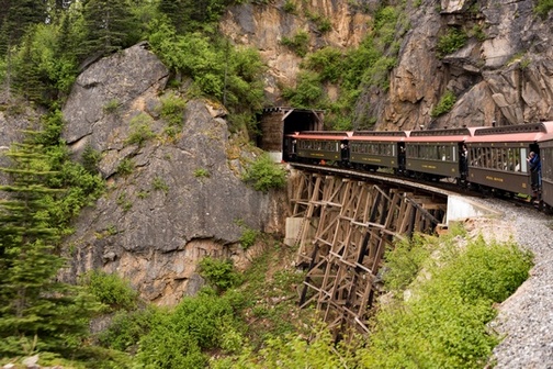 View of a section of a train crossing wooden trellis and entering tunnel into mountain.