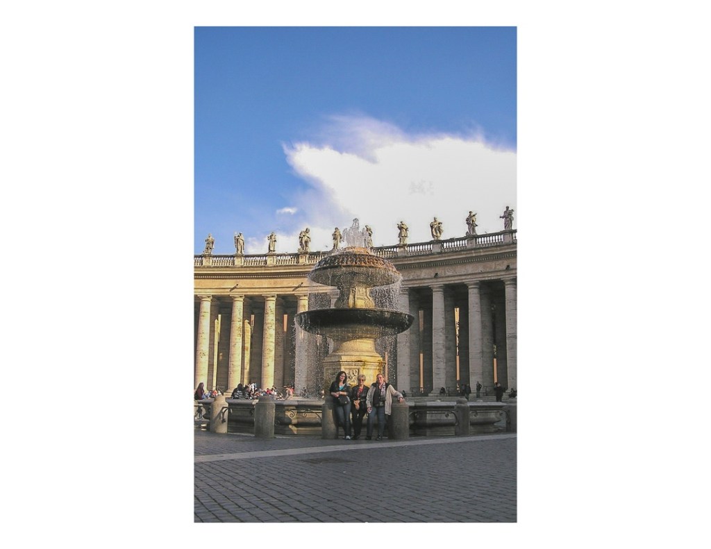 Three women standing in front of fountain, with colonnades and statues of saints in background, taken at Piazza San Pietro, Vatican City.