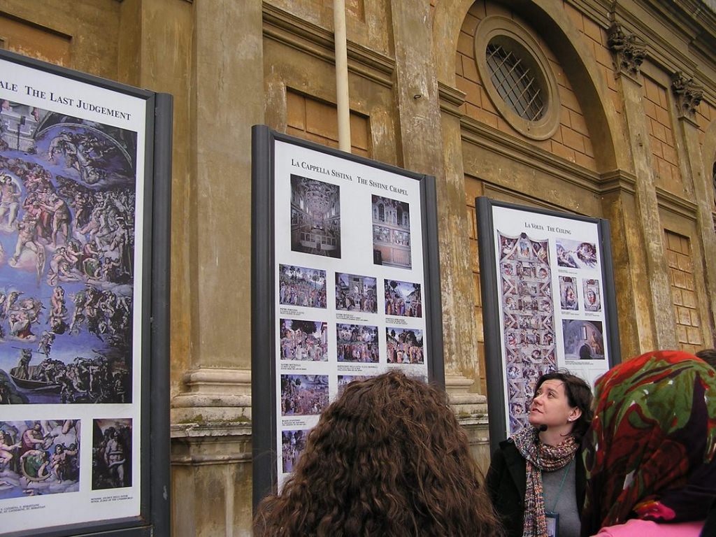 Female tour guide looking at and explaining large posters of information regarding the Sistine Chapel, with two female tourists in foreground.