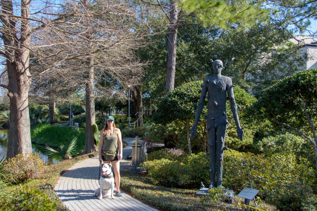 Woman and white dog on sidewalk adjacent to tall bronze sculpture in Besthoff Sculpture Garden, New Orleans.